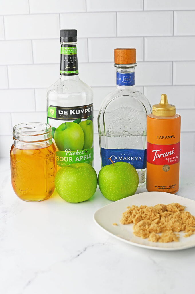 caramel apple margarita ingredients on a counter- apple juice, sour apple pucker, tequila, and Torani caramel sauce